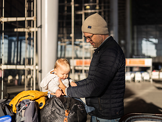 Image showing Fatherat comforting his tired infant baby boy child sitting on top of luggage cart in front of airport terminal station while traveling wih family.