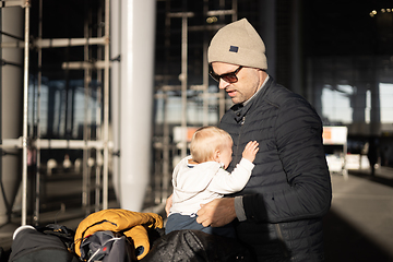 Image showing Fatherat comforting his crying infant baby boy child tired sitting on top of luggage cart in front of airport terminal station while traveling wih family.