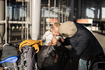 Image showing Fatherat comforting his crying infant baby boy child tired sitting on top of luggage cart in front of airport terminal station while traveling wih family.