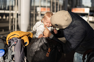 Image showing Fatherat comforting his crying infant baby boy child tired sitting on top of luggage cart in front of airport terminal station while traveling wih family.