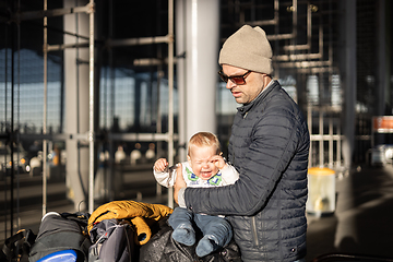 Image showing Fatherat comforting his crying infant baby boy child tired sitting on top of luggage cart in front of airport terminal station while traveling wih family.