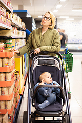 Image showing Casualy dressed mother choosing canned products in department of supermarket grocery store with her infant baby boy child in stroller.