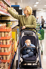 Image showing Casualy dressed mother choosing canned products in department of supermarket grocery store with her infant baby boy child in stroller.