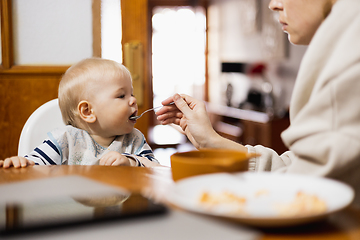 Image showing Mother spoon feeding her infant baby boy child sitting in high chair at the dining table in kitchen at home