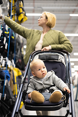 Image showing Casualy dressed mother choosing sporty shoes and clothes products in sports department of supermarket store with her infant baby boy child in stroller.