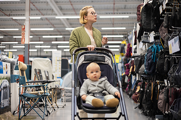 Image showing Casualy dressed mother choosing sporty shoes and clothes products in sports department of supermarket store with her infant baby boy child in stroller.