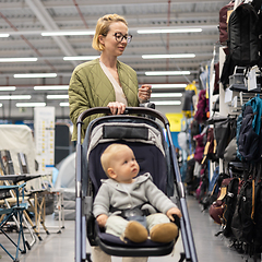 Image showing Casualy dressed mother choosing sporty shoes and clothes products in sports department of supermarket store with her infant baby boy child in stroller.