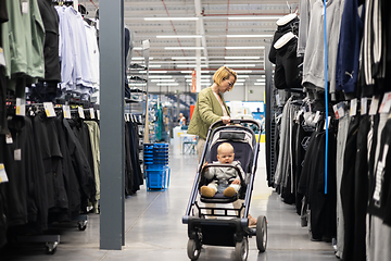 Image showing Casualy dressed mother choosing sporty shoes and clothes products in sports department of supermarket store with her infant baby boy child in stroller.