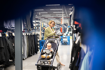 Image showing Casualy dressed mother choosing sporty shoes and clothes products in sports department of supermarket store with her infant baby boy child in stroller.