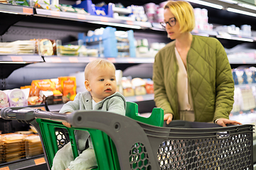 Image showing Casualy dressed mother choosing canned products in department of supermarket grocery store with her infant baby boy child in shoping cart