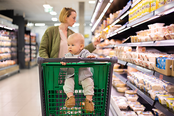 Image showing Mother pushing shopping cart with her infant baby boy child down department aisle in supermarket grocery store. Shopping with kids concept.