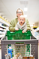 Image showing Mother pushing shopping cart with her infant baby boy child down department aisle in supermarket grocery store. Shopping with kids concept.