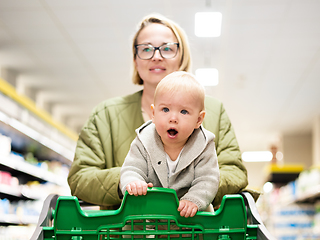 Image showing Mother pushing shopping cart with her infant baby boy child down department aisle in supermarket grocery store. Shopping with kids concept.