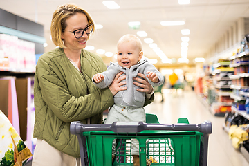 Image showing Mother pushing shopping cart with her infant baby boy child down department aisle in supermarket grocery store. Shopping with kids concept.