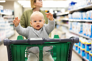 Image showing Mother pushing shopping cart with her infant baby boy child down department aisle in supermarket grocery store. Shopping with kids concept.