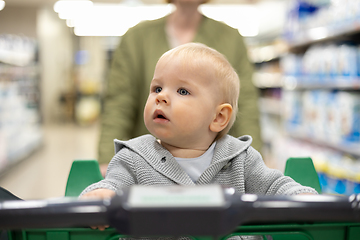 Image showing Mother pushing shopping cart with her infant baby boy child down department aisle in supermarket grocery store. Shopping with kids concept.