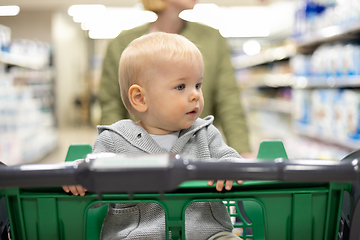 Image showing Mother pushing shopping cart with her infant baby boy child down department aisle in supermarket grocery store. Shopping with kids concept.