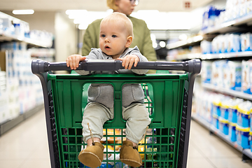 Image showing Mother pushing shopping cart with her infant baby boy child down department aisle in supermarket grocery store. Shopping with kids concept.