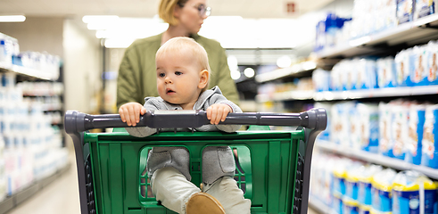 Image showing Mother pushing shopping cart with her infant baby boy child down department aisle in supermarket grocery store. Shopping with kids concept.