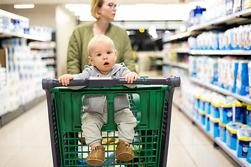 Image showing Mother pushing shopping cart with her infant baby boy child down department aisle in supermarket grocery store. Shopping with kids concept.