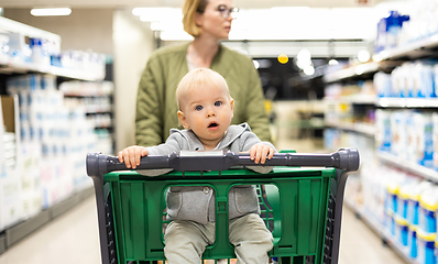 Image showing Mother pushing shopping cart with her infant baby boy child down department aisle in supermarket grocery store. Shopping with kids concept.