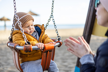 Image showing Mother pushing her infant baby boy child on a swing on sandy beach playground outdoors on nice sunny cold winter day in Malaga, Spain.