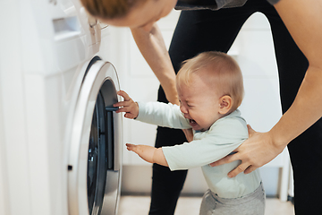 Image showing Little infant baby boy child demanding mother's attention while she is multitasking, trying to do some household chores in kitchen at home. Mother on maternity leave.