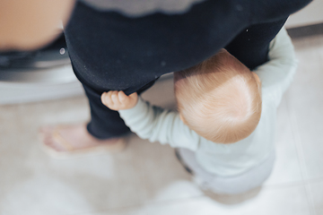 Image showing Top down view of little infant baby boy hiding between mothers legs demanding her attention while she is multitasking, trying to do some household chores in kitchen at home. Mother on maternity leave.