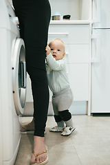 Image showing Little infant baby boy child hiding between mothers legs demanding her attention while she is multitasking, trying to do some household chores in kitchen at home. Mother on maternity leave.