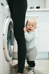 Image showing Little infant baby boy child hiding between mothers legs demanding her attention while she is multitasking, trying to do some household chores in kitchen at home. Mother on maternity leave.