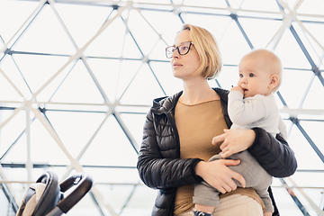 Image showing Mother carying his infant baby boy child, pushing stroller at airport departure terminal waiting at boarding gates to board an airplane. Family travel with baby concept.