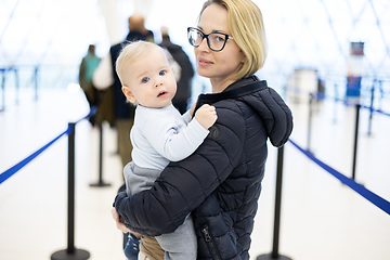 Image showing Mother carying his infant baby boy child queuing at airport terminal in passport control line at immigrations departure before moving to boarding gates to board an airplane. Travel with baby concept.