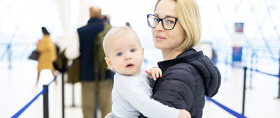 Image showing Mother carying his infant baby boy child queuing at airport terminal in passport control line at immigrations departure before moving to boarding gates to board an airplane. Travel with baby concept.