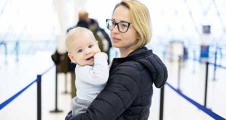 Image showing Mother carying his infant baby boy child queuing at airport terminal in passport control line at immigrations departure before moving to boarding gates to board an airplane. Travel with baby concept.