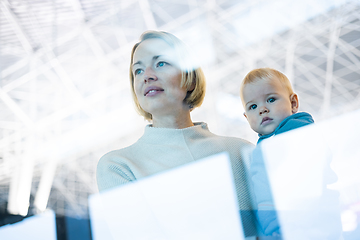 Image showing Thoughtful young mother looking trough window holding his infant baby boy child while waiting to board an airplane at airport terminal departure gates. Travel with baby concept.