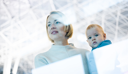 Image showing Thoughtful young mother looking trough window holding his infant baby boy child while waiting to board an airplane at airport terminal departure gates. Travel with baby concept.