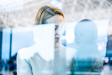 Image showing Thoughtful young mother looking trough window holding his infant baby boy child while waiting to board an airplane at airport terminal departure gates. Travel with baby concept.