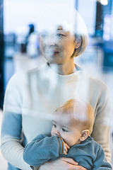 Image showing Thoughtful young mother looking trough window holding his infant baby boy child while waiting to board an airplane at airport terminal departure gates. Travel with baby concept.