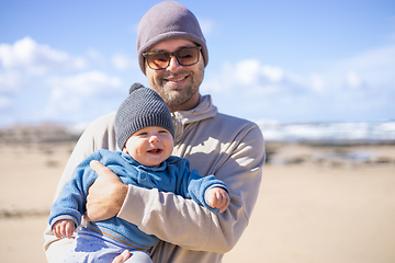 Image showing Father enjoying pure nature holding and playing with his infant baby boy son in on windy sandy beach of Famara, Lanzarote island, Spain. Family travel and parenting concept.