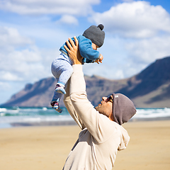 Image showing Father enjoying pure nature holding and playing with his infant baby boy son in on windy sandy beach of Famara, Lanzarote island, Spain. Family travel and parenting concept.