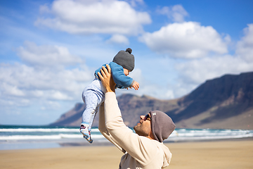 Image showing Father enjoying pure nature holding and playing with his infant baby boy son in on windy sandy beach of Famara, Lanzarote island, Spain. Family travel and parenting concept.