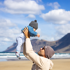 Image showing Father enjoying pure nature holding and playing with his infant baby boy son in on windy sandy beach of Famara, Lanzarote island, Spain. Family travel and parenting concept.