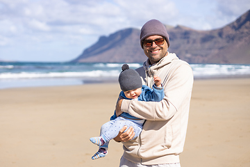 Image showing Father enjoying pure nature holding and playing with his infant baby boy son in on windy sandy beach of Famara, Lanzarote island, Spain. Family travel and parenting concept.