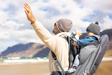 Image showing Young father rising hands to the sky while enjoying pure nature carrying his infant baby boy son in backpack on windy sandy beach. Family travel concept.