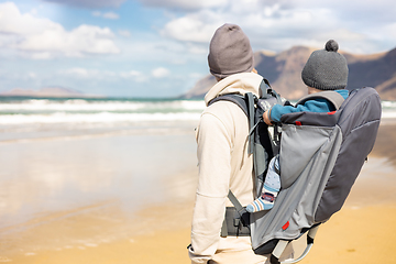 Image showing Young father carrying his infant baby boy son in backpack on windy sandy beach. Family travel and winter vacation concept.