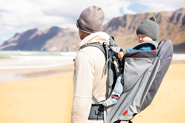 Image showing Young father carrying his infant baby boy son in backpack on windy sandy beach. Family travel and winter vacation concept.