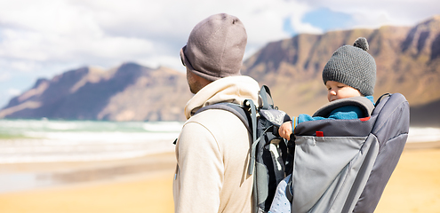 Image showing Young father carrying his infant baby boy in backpack on windy sandy beach. Family travel and vacation concept.