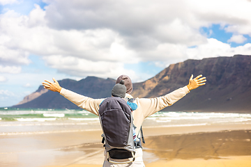 Image showing Young father rising hands to the sky while enjoying pure nature carrying his infant baby boy son in backpack on windy sandy beach of Famara, Lanzarote island, Spain. Family travel concept.