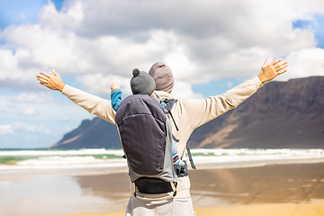 Image showing Young father rising hands to the sky while enjoying pure nature carrying his infant baby boy son in backpack on windy sandy beach of Famara, Lanzarote island, Spain. Family travel concept.