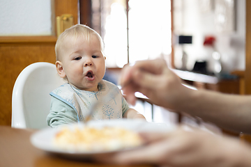 Image showing Mother spoon feeding her infant baby boy child sitting in high chair at the dining table in kitchen at home